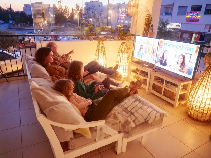 A modern Israeli living room in the evening, warmly lit, cozy atmosphere, a young Israeli family (parents in their 30s, two children ages 6-10) sitting together on a comfortable sofa, watching TV, natural and realistic faces, proportional facial features, soft and relaxed expressions, cinematic lighting, realistic details, laptop on side table showing yoga or meditation app, large window showing a quiet city street with closed restaurants and empty sidewalks, ultra-detailed, high-resolution, natural color tones --chaos 25 --ar 4:3 --stylize 450 --weird 300 --v 7 Job ID: cf002ca0-3be8-4ea5-9512-639a950c7814